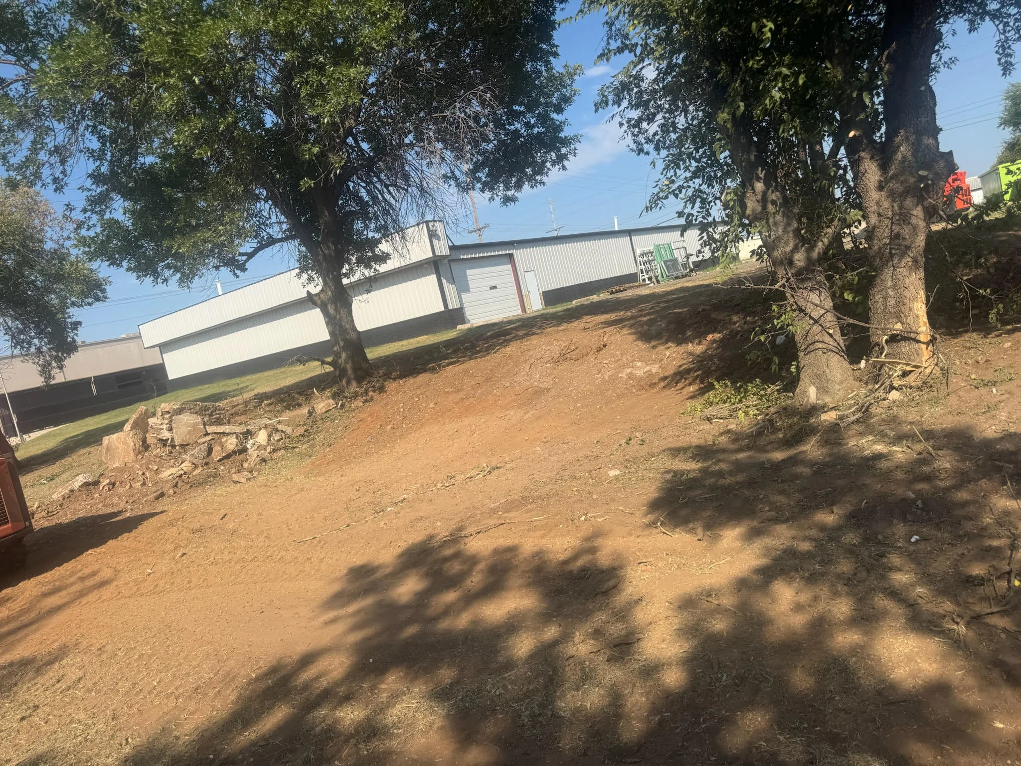 Graded dirt lot with mature trees and metal building in background