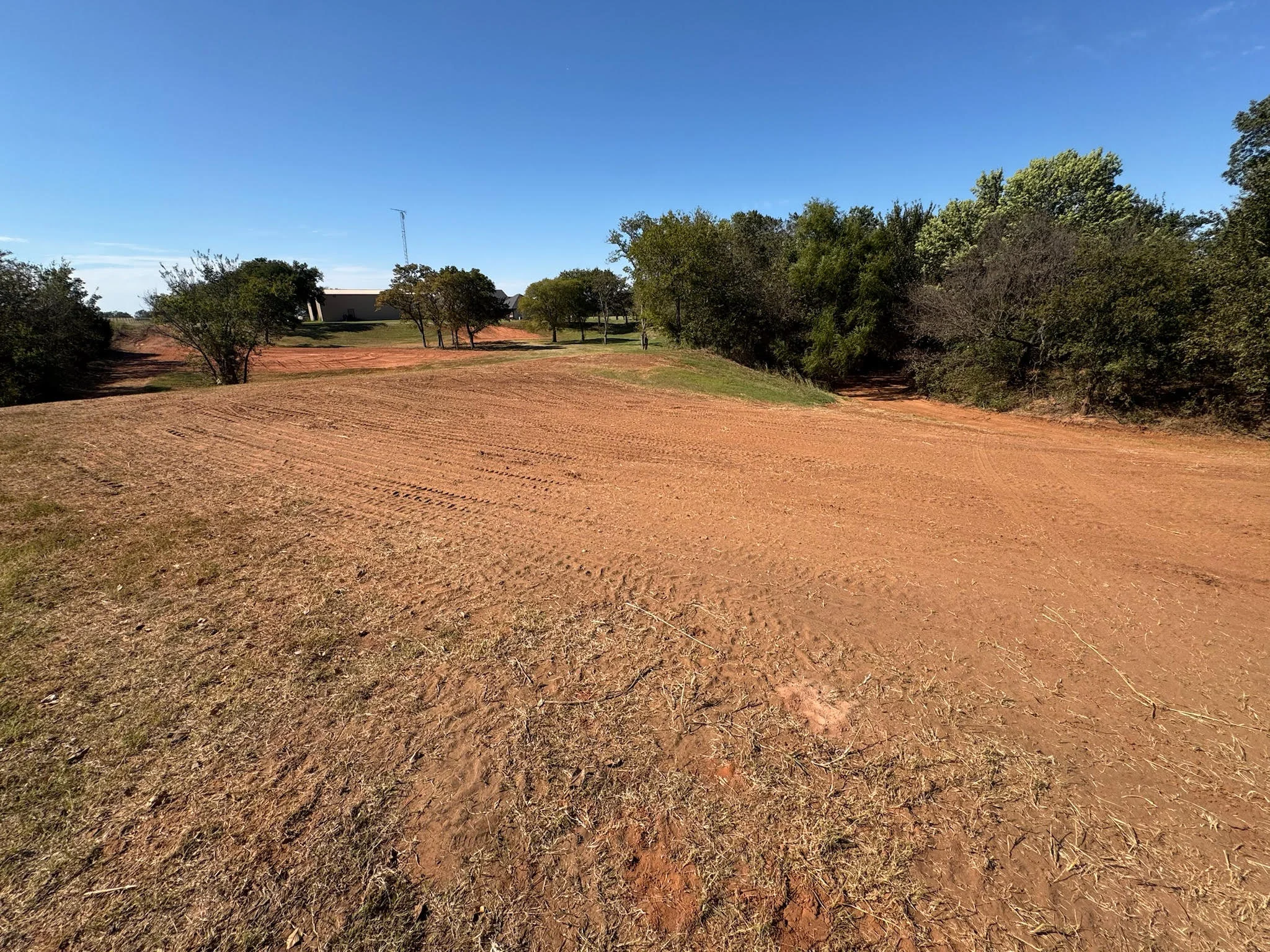 Excavated residential lot with red clay soil, cleared building area, and mature trees