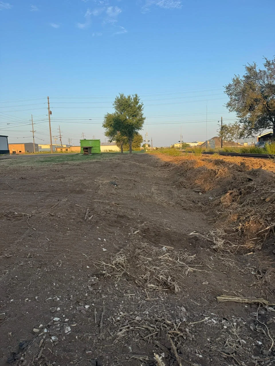 Excavated commercial lot with graded earth, utility poles, and industrial buildings in background