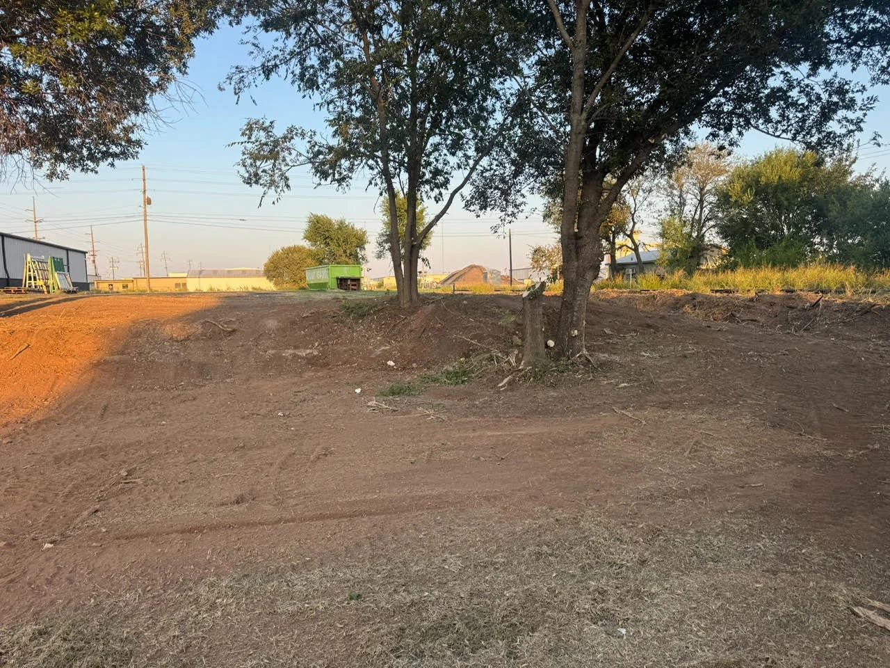 Excavated commercial lot with graded soil, mature trees, and equipment staging area