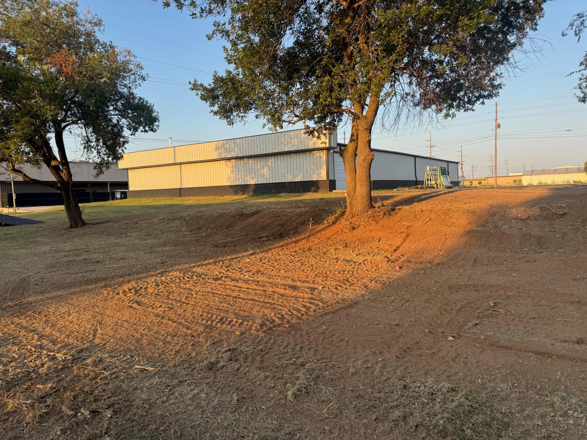 Graded dirt lot with metal warehouse buildings and mature oak tree