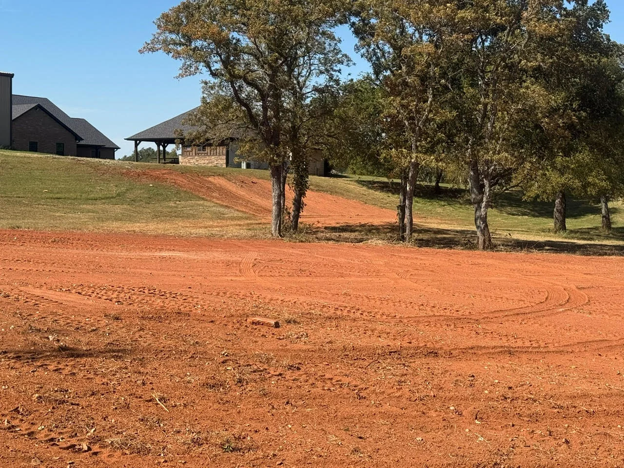 Red clay driveway excavation with graded surface and mature oak trees on rural property